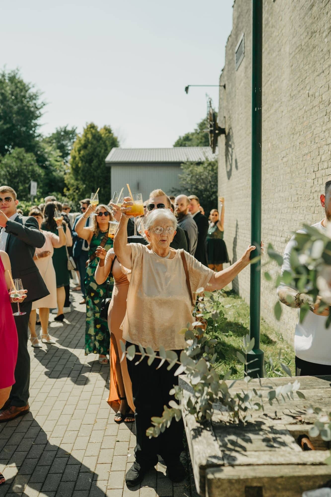 Dennis’ Oma hebt auf der Hochzeit lachend ihr Glas, hinter ihr stoßen die Gäste an